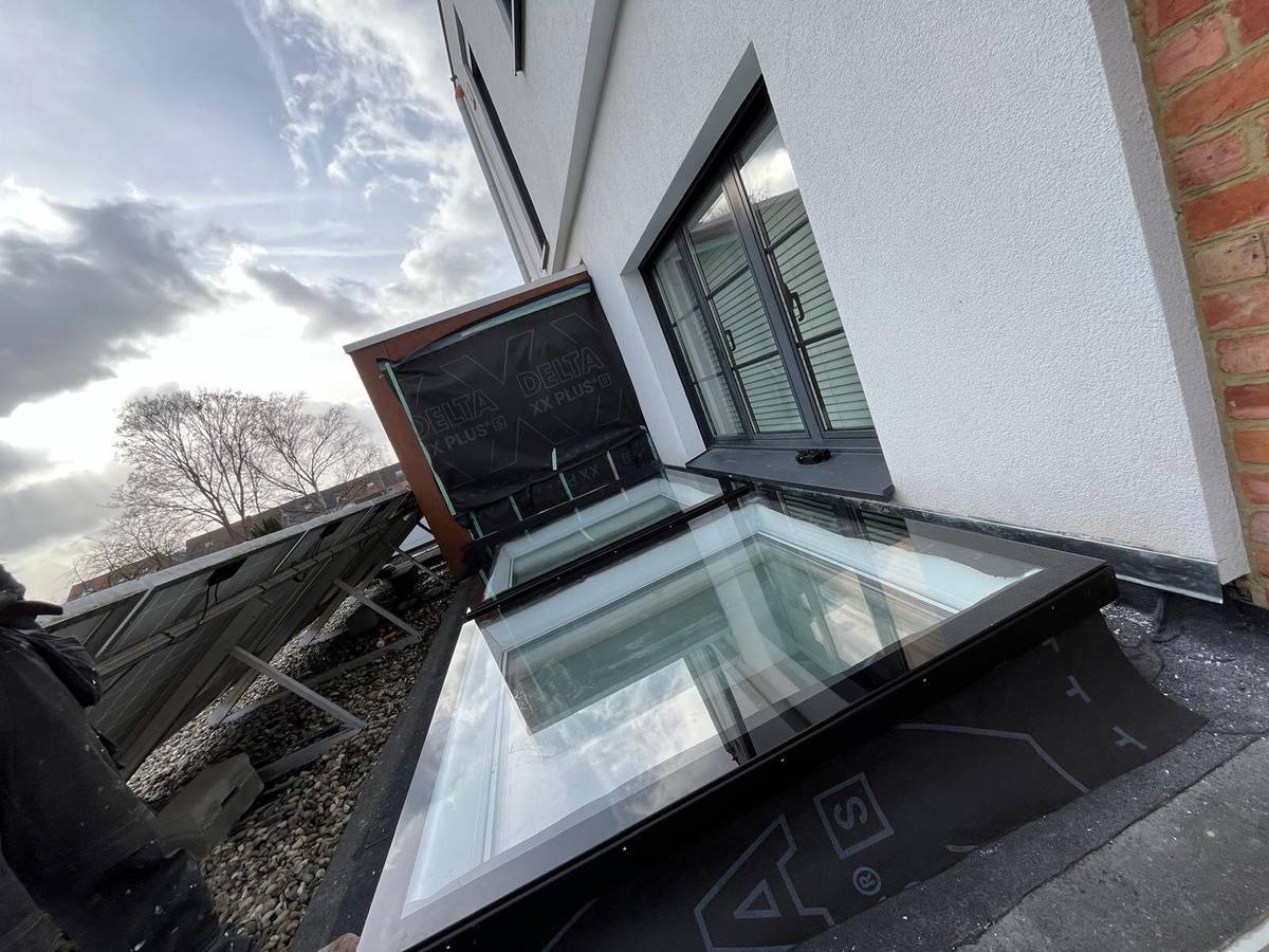 Overzicht van geïnstalleerde daklichten op een plat dak naast een wit huis, met zonnepanelen zichtbaar.