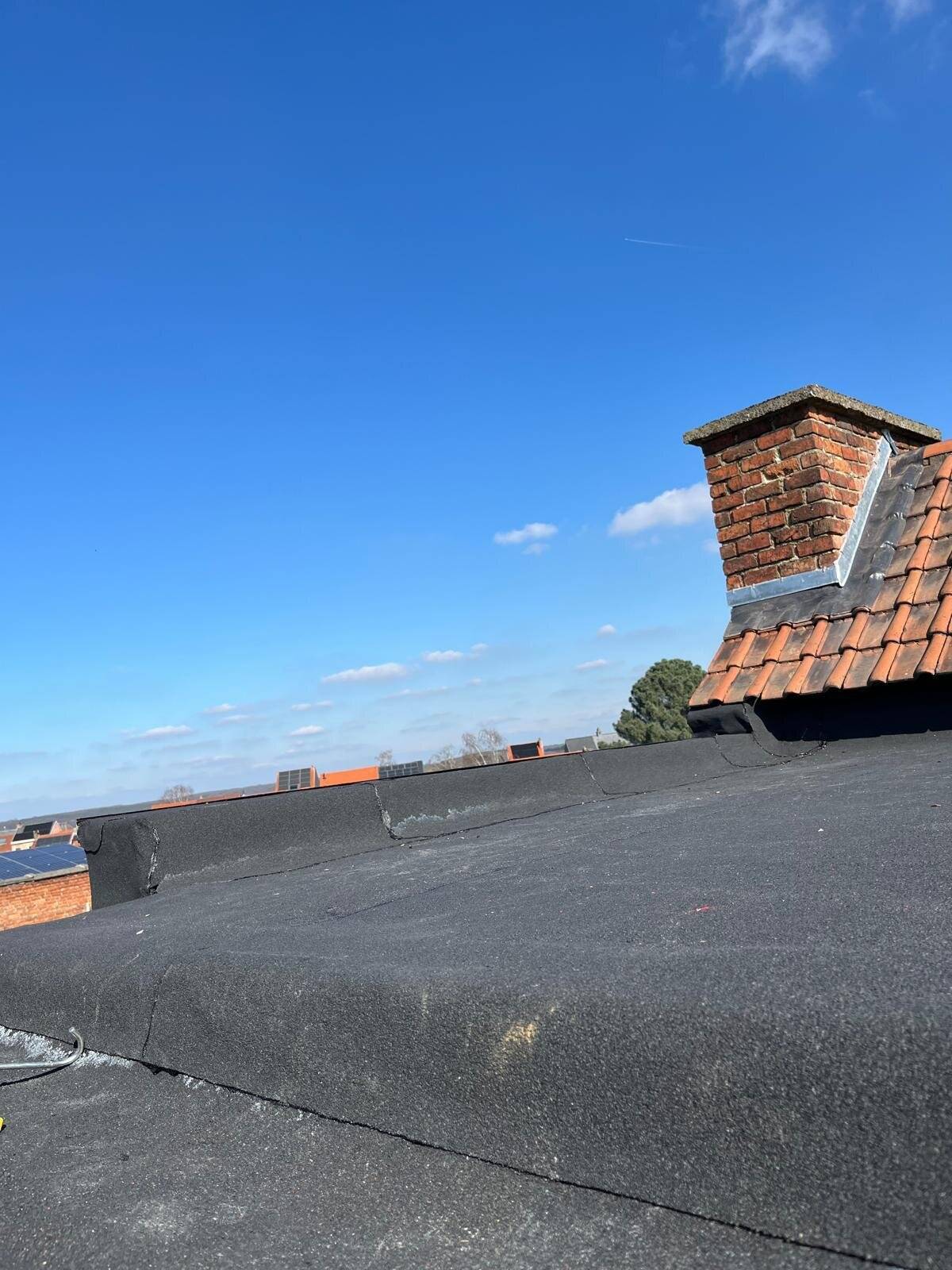 Zicht op een plat dak van bitumen naast een hellend dak met schoorsteen onder een blauwe lucht.