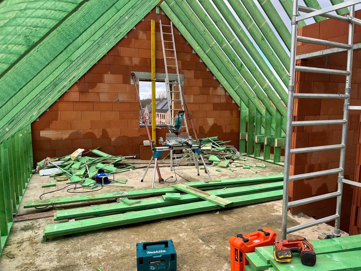 Binnenkant van een nieuw hellend dak in aanbouw met groene houten spanten en bakstenen muren.